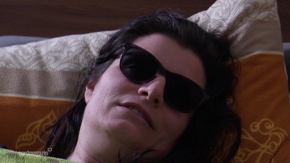 Eine Frau liegt mit dunkler Sonnenbrille im Bett. © Screenshot 